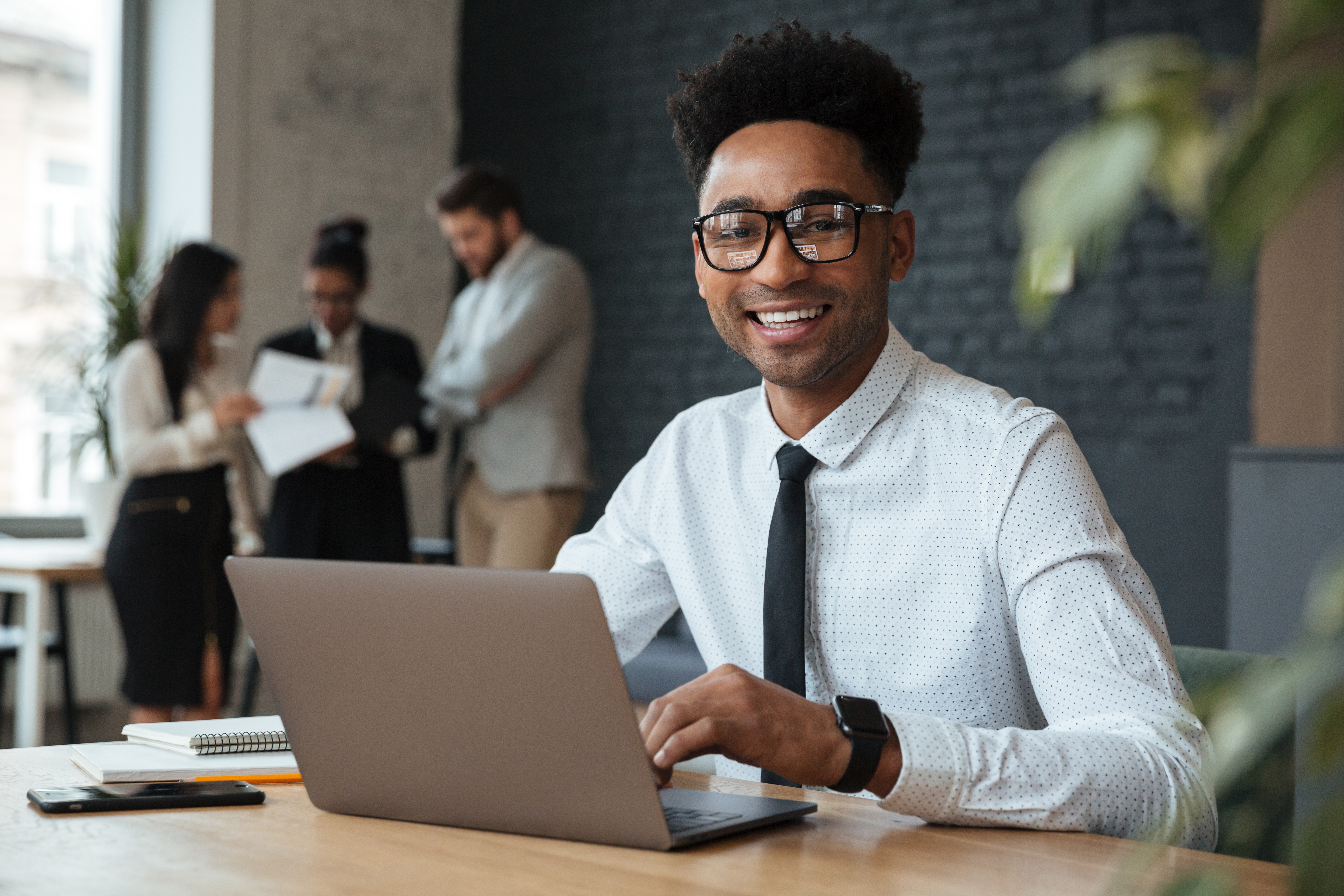 Photo of happy young african businessman sitting indoors using laptop computer. Looking camera.